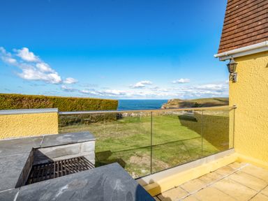 A patio with a built-in barbecue grill and glass railing overlooking a grassy area and ocean at Trevan House Lundy in Polzeath