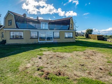 A yellow house with a glass balcony and large windows surrounded by a lawn with patches of dry grass at Trevan House Lundy in Polzeath