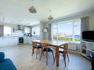 A kitchen and dining area with wooden table and chairs near large window with sea view at Windyhill in Polzeath