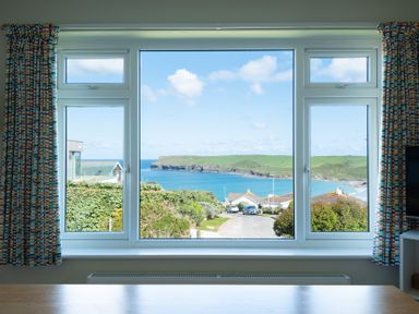 A window with colorful curtains showing a road leading to houses and a view of the sea and green hills at Windyhill in Polzeath