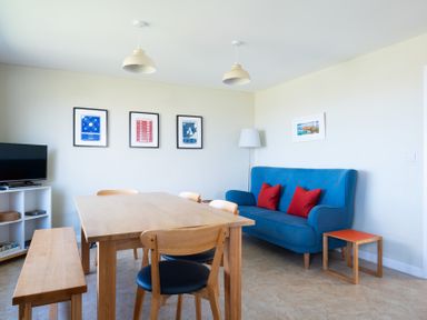 A dining area with a wooden table, chairs, bench, blue sofa with red pillows, TV on a white shelf, and wall art at Windyhill in Polzeath