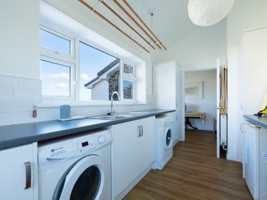 A laundry room with washing machines a sink white cabinets and a large window at Windyhill in Polzeath