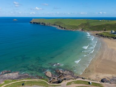 A beach with waves and surfers in the water with green cliffs and a clear blue sky at Windyhill in Polzeath