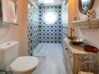 A bathroom with a toilet wooden vanity with a vessel sink patterned tiled shower area and round window at Trevanion in Rock