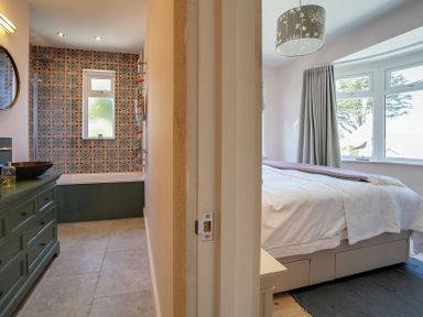 A bathroom with patterned tiles, bathtub, sink, and a bedroom with a bed, nightstand, and window at Trevanion in Rock