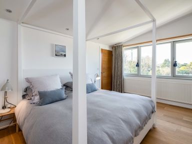 A bedroom with a four-poster bed grey bedding and pillows a wooden door and three windows with curtains at Seastar in Rock