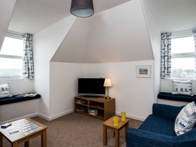A small sitting room with a television on a wooden stand cushions on window seats and two small tables with drinks at Pinewood 14 in Polzeath
