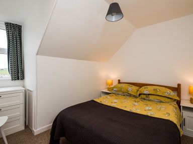 A bedroom with a bed covered with yellow floral bedding two bedside tables with lamps a chest of drawers and a window with patterned curtains at Pinewood 14 Polzeath