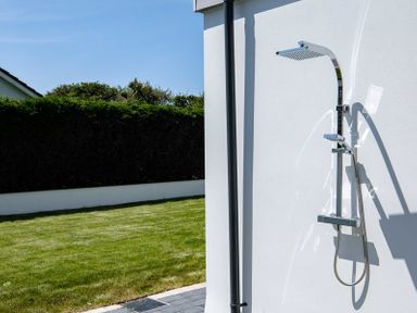 An outdoor shower attached to a white wall with a grassy yard and hedge in the background at Artemis Rock