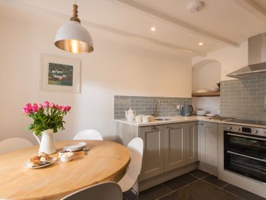 A kitchen with grey cabinets a wooden round dining table with flowers and plates at Kipper Cottage in Port Isaac