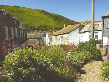 A garden with flowering plants and stone houses with a green hill in the background at Kipper Cottage Port Isaac
