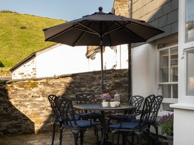 An outdoor patio with a round table and six chairs under a black umbrella at Kipper Cottage in Port Isaac