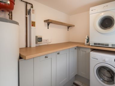 A utility room with a washing machine a dryer an iron wooden cabinets and a wooden wall shelf at Kipper Cottage in Port Isaac