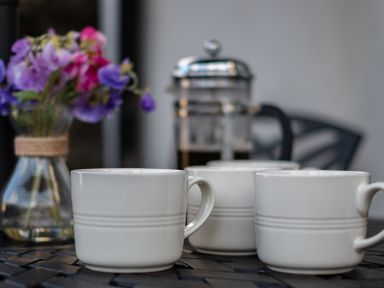 Three white coffee cups on a table with a flower vase and coffee press in the background at Kipper Cottage in Port Isaac