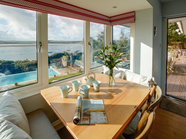 A dining area with a wooden table set with cups a teapot flowers and a magazine next to large windows overlooking a pool and water at Camel Point in Rock