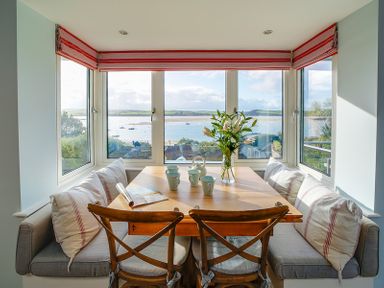 A dining nook with a wooden table four cups a vase with flowers two wooden chairs and cushioned bench seating by windows overlooking a waterfront at Camel Point in Rock