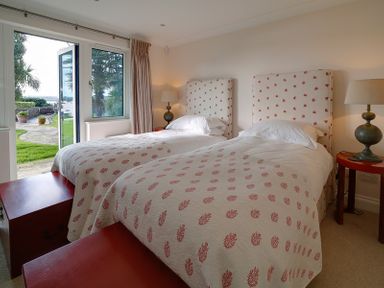 A bedroom with two single beds side by side covered in white quilts with red patterns with two bedside tables and lamps and a door opening to a patio and garden at Camel Point in Rock