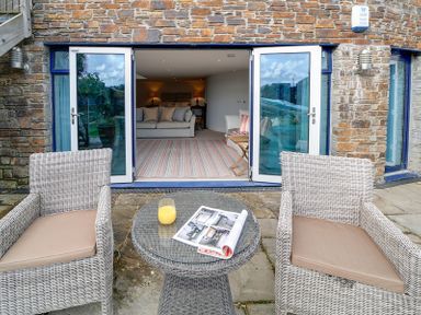 An outdoor seating area with two wicker chairs and a round table with a magazine and glass of orange juice in front of open doors leading to a living room with sofas and lamps at Camel Point in Rock