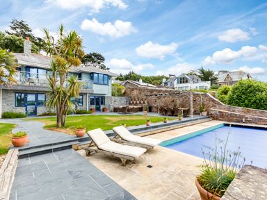 An outdoor area with two lounge chairs by a covered swimming pool a stone patio a lawn with a tree multiple potted plants and a modern house with large windows at Camel Point in Rock