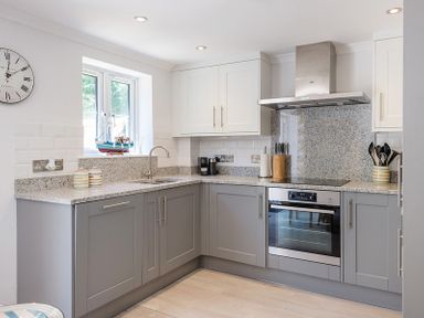 A kitchen with grey lower cabinets white upper cabinets a built-in oven and a window with a model boat on the sill at Stop Tide in Port Isaac