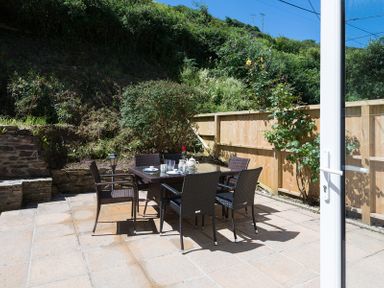 An outdoor patio with a glass table and six woven chairs surrounded by plants and a wooden fence at Stop Tide in Port Isaac