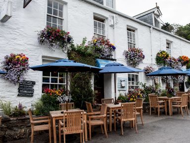 Outdoor seating area with wooden tables and chairs under blue umbrellas outside a white building with hanging flower baskets at Stop Tide in Port Isaac