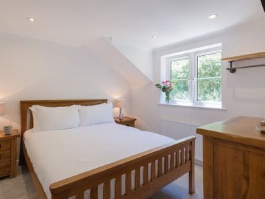 A bedroom with a wooden bed frame white bedding two bedside tables with lamps a window with flowers on the sill and a wooden dresser at Stop Tide in Port Isaac