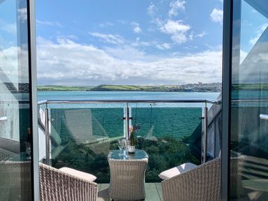 A balcony with two wicker chairs a small glass table with a wine bottle and glasses overlooking water and hills at Upper Deck in Rock
