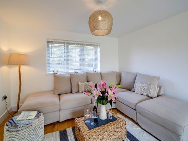 A living room with a beige sectional sofa a woven coffee table with flowers and wine glasses and a floor lamp near the window at Elmwood in Port Isaac