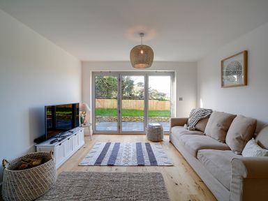 A living room with a beige sofa a TV on a white stand a lamp a basket and glass doors opening to a garden at Elmwood in Port Isaac