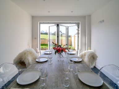 A dining room with a wooden table set with plates and glasses and transparent chairs with white fur throws at Elmwood in Port Isaac