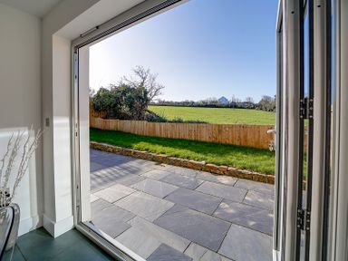 A view of a stone patio and grass fenced yard opening to a green field with trees and a house in the distance at Elmwood in Port Isaac