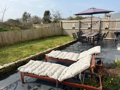 An outdoor patio with two cushioned lounge chairs a dining table with black chairs and an umbrella a barbecue grill and a lawn surrounded by wooden fencing at Elmwood in Port Isaac