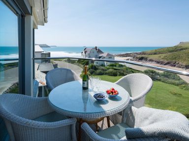 A balcony with a glass table and wicker chairs overlooking the ocean and a grassy area at Egloskerry in New Polzeath