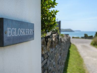 A sign reading Egloskerry on a white wall with a stone wall and a coastal road leading to the sea at Egloskerry in New Polzeath