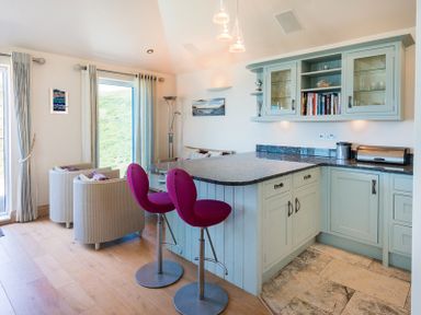 A kitchen with light blue cabinets a granite countertop two pink bar stools and two beige armchairs at Egloskerry in New Polzeath