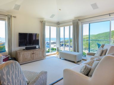 A living room with sofas a television and large windows overlooking the ocean and hills at Egloskerry in New Polzeath