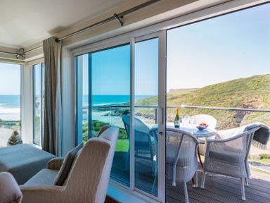 A seating area with armchairs and a table with a bottle and glasses on a balcony overlooking the ocean and hills at Egloskerry in New Polzeath