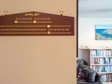 A wall-mounted Riley scoreboard with sliding markers and a glimpse of a living room with a sofa and a bookshelf at Egloskerry in New Polzeath