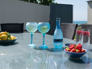 A glass table with blue glasses, a blue bottle, bowls of lemons and strawberries, and two glasses with raspberries outdoors at Egloskerry in New Polzeath
