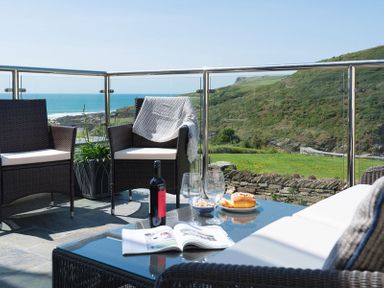 A patio with wicker chairs a glass table bottle of wine two glasses and a view of hills and ocean at Egloskerry in New Polzeath
