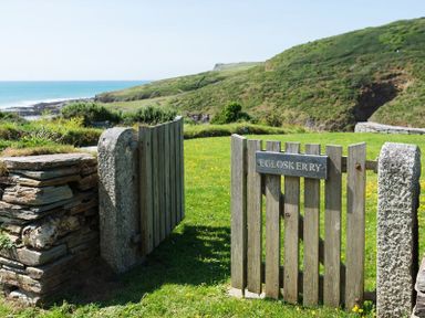 A wooden gate with a stone post and a sign reading Egioskerry with a grassy field and ocean in the background at Egloskerry in New Polzeath