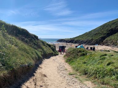 A sandy path between grassy hills leading to a beach with people and tents at Egloskerry in New Polzeath