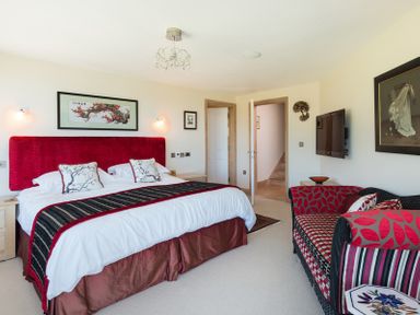 A bedroom with a large bed with red headboard and decorative pillows a patterned sofa a wall-mounted tv and framed artwork at Egloskerry in New Polzeath