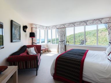A bedroom with a bed a red and black patterned sofa large windows with floral curtains and a view of hills at Egloskerry in New Polzeath