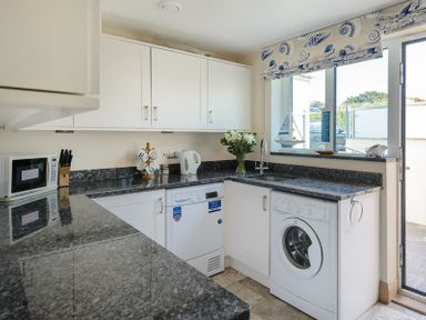 A kitchen with marble countertops white cabinets appliances and a window with a sea-themed valance at Egloskerry in New Polzeath