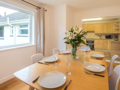 A dining table set with plates knives and wine glasses and a vase of flowers in a room with a window and a view into a kitchen at Greenaway 4 Rockhaven Gardens Rock
