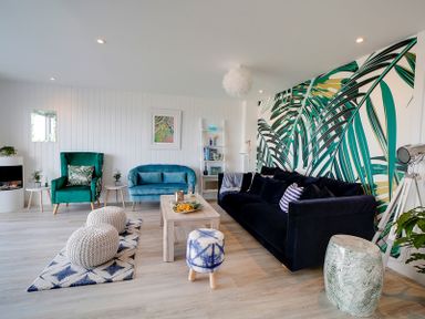A living room with a green armchair blue loveseat black sofa large leaf patterned wallpaper wooden coffee table knitted poufs and patterned stool at Seahaven in Port Isaac