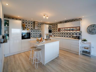 A modern kitchen with white cabinets a central island with two stools tiled backsplash and a large wall clock at Seahaven in Port Isaac