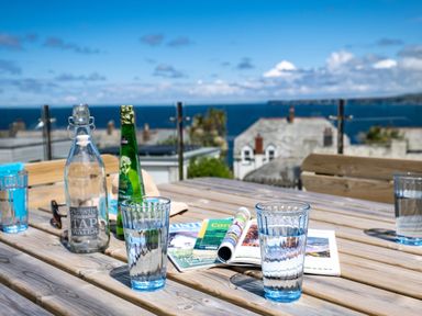 A wooden table with glasses of water a water bottle a green bottle and magazines outdoors with a sea view at Seahaven in Port Isaac